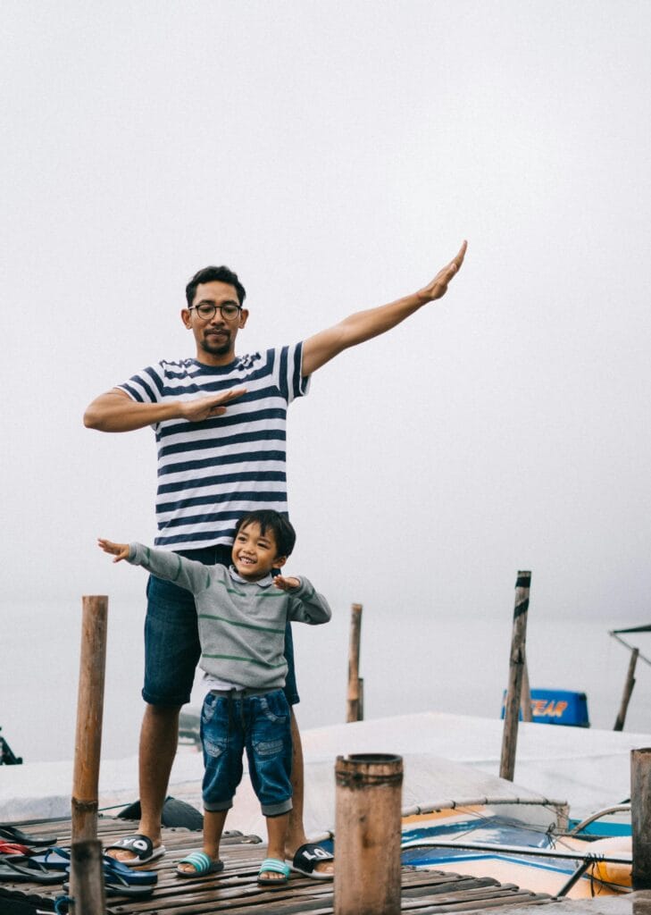 Father and son posing playfully on a wooden dock by the water, enjoying a day outdoors.
