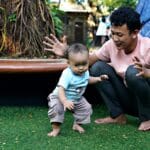A father encourages his baby to take first steps on a sunny day in a park, barefoot on green grass.