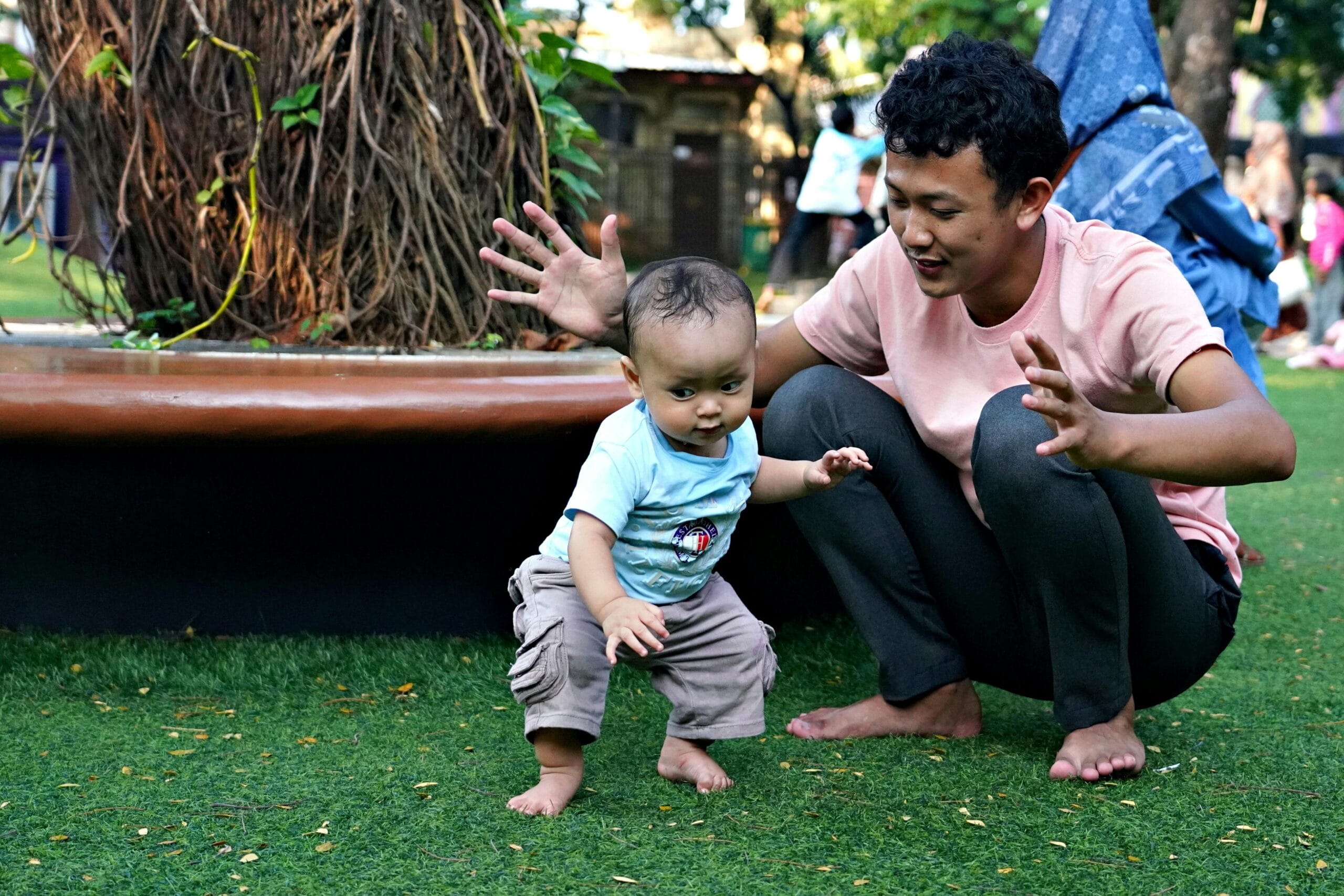 A father encourages his baby to take first steps on a sunny day in a park, barefoot on green grass.