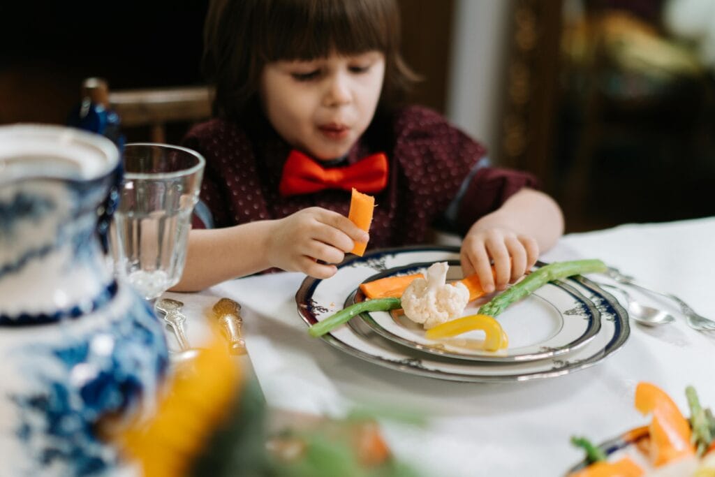 A child in a red bow tie enjoys a healthy meal with vegetables indoors.