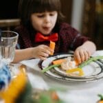A child in a red bow tie enjoys a healthy meal with vegetables indoors.