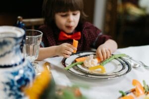 A child in a red bow tie enjoys a healthy meal with vegetables indoors.